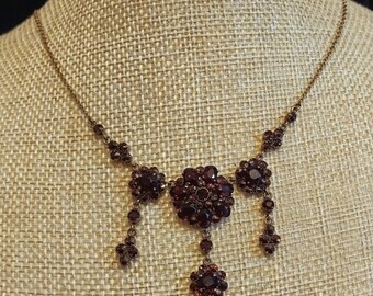 May include: A vintage-style necklace featuring a large, ornate pendant crafted from dark red glass stones. The pendant is surrounded by smaller, similar stones and hangs from a delicate chain.