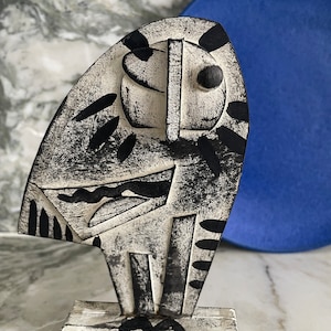 Puede incluir: Una escultura de madera abstracta en blanco y negro de una figura estilizada con formas geométricas. La escultura está sobre una base de madera y está delante de un fondo azul.
