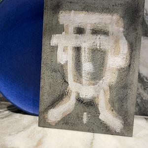 Op de afbeelding: Abstract kunstwerk op een rechthoekig, grijs canvas met een wit en beige geschilderd ontwerp. Een grote, ronde, blauwe plaat is gedeeltelijk zichtbaar aan de linkerkant. Het kunstwerk heeft een textuur. De plaat heeft een diameter van ongeveer 25 cm.