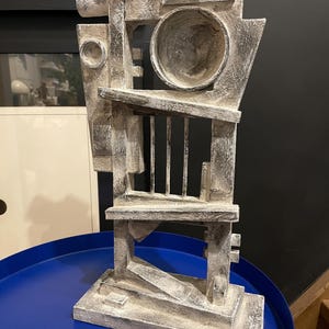 Puede incluir: Una escultura abstracta de madera de color blanco y gris. La escultura está compuesta por formas geométricas y tiene una superficie rugosa y texturizada.