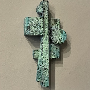 Könnte beinhalten: Abstrakte Wandskulptur aus Metall mit einer grün-blauen Patina. Die Skulptur besteht aus geometrischen Formen, darunter Rechtecke und Kreise.