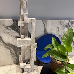 Può includere: Una scultura verticale alta composta da blocchi rettangolari e pilastri interconnessi. L'oggetto presenta una finitura strutturata e usurata nei toniata in tonalità bianco sporco, beige e grigio chiaro, creando una struttura geometrica a strati.