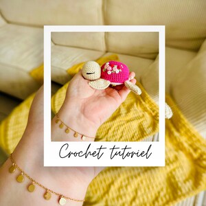 Könnte beinhalten: Eine gehäkelte rosa und weiße Schildkröte mit Blumendetails. Die Schildkröte wird in einer Hand mit einem goldenen Armband gehalten. Das Bild hat den Text "Crochet tutorial" unten.