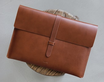 May include: A brown leather laptop sleeve with a flap closure and a strap. The sleeve has the word "PAUSE" embossed on the front.