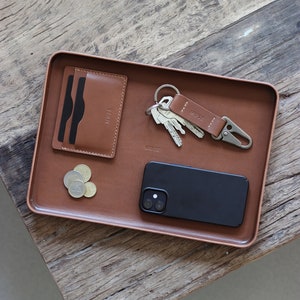 Op de afbeelding: Een bruine leren dienblad met een zwarte telefoon, een leren kaarthouder met het woord "MUSE" erop, een sleutelset met een leren sleutelhanger waarop "MUSE" staat, en een paar munten.