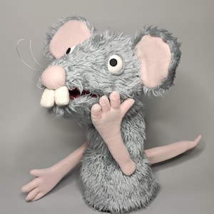 Peut inclure: Marionnette de souris grise en peluche avec des oreilles, un nez et des mains roses. La marionnette a de grandes dents blanches, une bouche grande ouverte et de longues moustaches. Les bras de la souris sont tendus et la marionnette semble surprise.