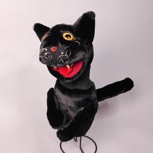 May include: Black plush cat puppet with a red mouth and yellow eyes. The puppet is on a metal stand.