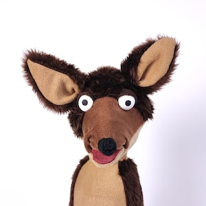 May include: A brown and tan puppet with large white eyes and a red mouth. The puppet is on a black stand.