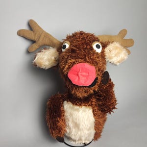 May include: A brown and white plush reindeer puppet with a red nose and large, white eyes. The puppet is wearing a brown felt hat and has a black felt mouth.