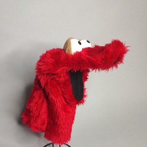 May include: A plush red character head cover with white eyes, a tan nose, and a black mouth. The cover is made of soft, fuzzy material and is designed to fit over a golf club head. The cover is designed to look like a popular children's character.