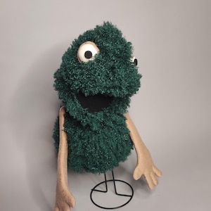 May include: A handmade puppet with a green, textured body and large, white-and-black eyes. The puppet has tan arms and hands, and a black mouth. It is mounted on a black wire stand, perfect for display or play.