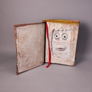 Puede incluir: Un libro abierto con un marcador de cinta roja. El libro tiene una cubierta de papel texturizado y un rostro esculpido en arcilla blanca es visible dentro del libro.