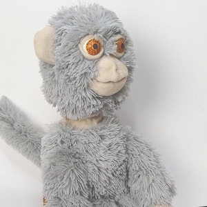 Può includere: Un peluche di scimmia grigia con grandi occhi espressivi e un sorriso amichevole. Il giocattolo ha una consistenza morbida e pelosa e orecchie e viso color beige. Le braccia della scimmia sono tese e sembra salutare.