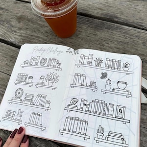 May include: An open notebook with a reading challenge theme. The notebook has a grid pattern and is filled with black and white illustrations of bookshelves with books, plants, and other decorative items. The text "Reading Challenge" is written at the top of the page.