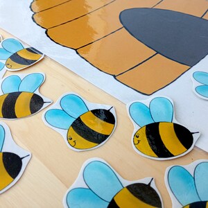 Puede incluir: Un conjunto de pegatinas de abejas amarillas y negras con alas azules. Las abejas están dispuestas en línea sobre una superficie de madera. Hay una colmena dibujada en una hoja de papel blanca en el fondo.