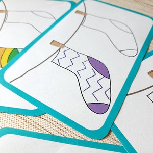 May include: Four white cards with a turquoise border, each featuring a sock hanging on a clothesline with a clothespin. The socks are decorated with different patterns: a rainbow, a chevron pattern, a purple zig-zag, and stars.