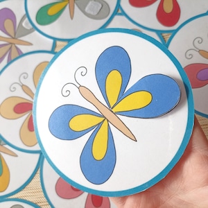 May include: A set of colorful paper butterfly cutouts with blue, yellow, red, green, and purple accents. The butterflies are on a white background with a teal border.
