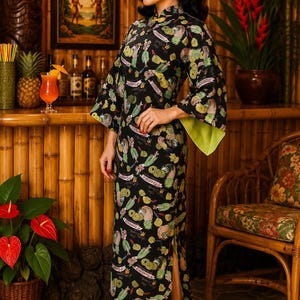 May include: A black floor-length dress featuring a mandarin collar, three-quarter length sleeves with lime green cuffs, and a side slit. The fabric is printed with a repeating pattern of pineapples, tropical foliage, and pink ribbon banners.