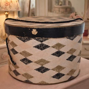 May include: A round, vintage hat box with a black rim and lid, featuring a repeating pattern of black and gold diamonds on a cream background. The box has a black rope handle and sits on a pink surface.