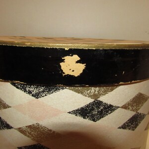 May include: An oval-shaped vintage hat box with a black rim and a gold-colored trim. The box features a repeating diamond pattern in black, gold, and pink on a cream background. A dark blue rope handle is visible.