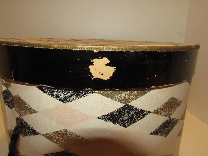 May include: An oval-shaped vintage hat box with a black rim and a gold-colored trim. The box features a repeating diamond pattern in black, gold, and pink on a cream background. A dark blue rope handle is visible.