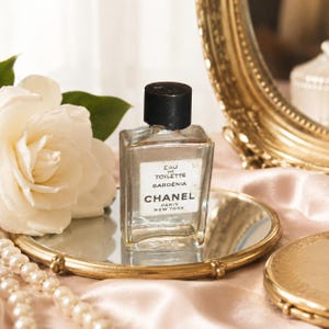 May include: A clear glass perfume bottle with a black cap and a white label reading EAU DE TOILETTE GARDENIA CHANEL PARIS NEW YORK. The bottle is positioned on a small round mirror next to a large white flower and a string of white pearls.