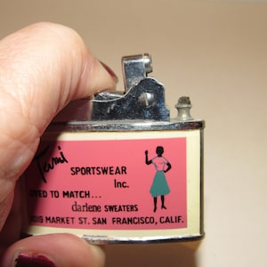 May include: Vintage metal lighter with a pink and cream label. The label features the text "Tami SPORTSWEAR Inc." and an illustration of a woman in a skirt. The lighter is being held by a hand.