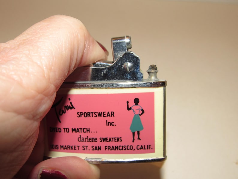 May include: Vintage metal lighter with a pink and cream label. The label features the text "Tami SPORTSWEAR Inc." and an illustration of a woman in a skirt. The lighter is being held by a hand.
