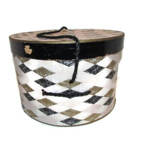 May include: A vintage round hat box with a black and white diamond pattern. The lid and base are trimmed in black, with a gold accent. A black rope handle is attached to the top. The box is likely made of cardboard or a similar material.
