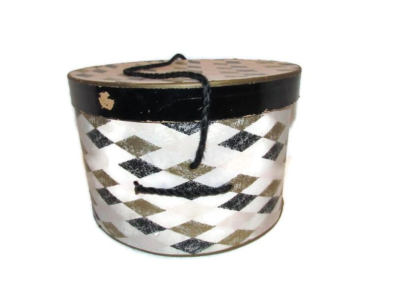 May include: A vintage round hat box with a black and white diamond pattern. The lid and base are trimmed in black, with a gold accent. A black rope handle is attached to the top. The box is likely made of cardboard or a similar material.