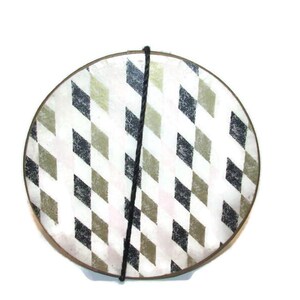 May include: Round decorative plate with a geometric diamond pattern in black, olive green, and white. A black cord is attached to the top of the plate. The plate has a brown rim.