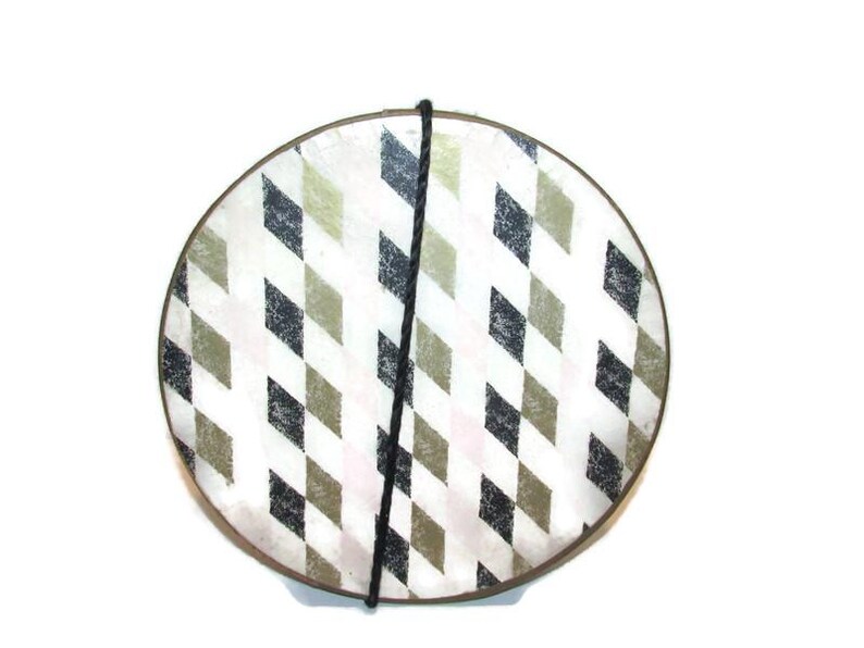 May include: Round decorative plate with a geometric diamond pattern in black, olive green, and white. A black cord is attached to the top of the plate. The plate has a brown rim.