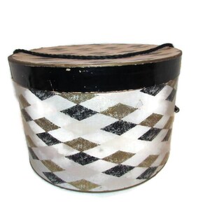 May include: A vintage round hat box with a black top and a white and diamond pattern. The box has a black rope handle and is likely made of cardboard or a similar material. The box is a decorative storage item.