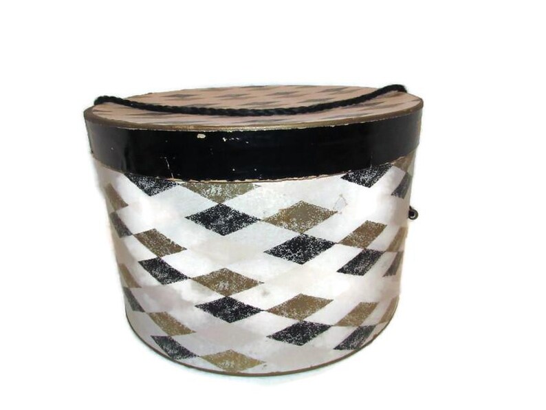 May include: A vintage round hat box with a black top and a white and diamond pattern. The box has a black rope handle and is likely made of cardboard or a similar material. The box is a decorative storage item.