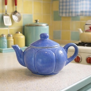 Puede incluir: Una tetera de cerámica azul con un cuerpo redondeado y una tapa, colocada sobre una encimera de cocina. La tetera tiene un asa y un pico. Utensilios de cocina y otros artículos de cocina son visibles en el fondo.