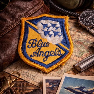 May include: A vintage-themed composition featuring a Blue Angels patch with a yellow and blue shield design, a leather aviator bag, sunglasses, and a watch. The patch has the words "Blue Angels" in gold lettering.