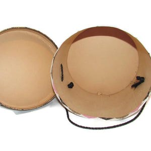 May include: An open, round, cardboard box with a lid. The interior is tan, and the exterior is decorated with a floral pattern. A black cord is attached to the side of the box.