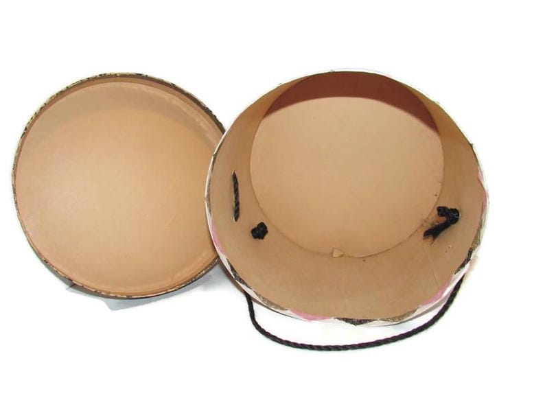 May include: An open, round, cardboard box with a lid. The interior is tan, and the exterior is decorated with a floral pattern. A black cord is attached to the side of the box.