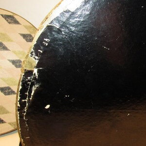 May include: Close-up of two round vintage hat boxes. One box is black with a glossy finish, showing signs of wear and tear. The other box has a cream and tan diamond pattern. The boxes are likely made of cardboard.