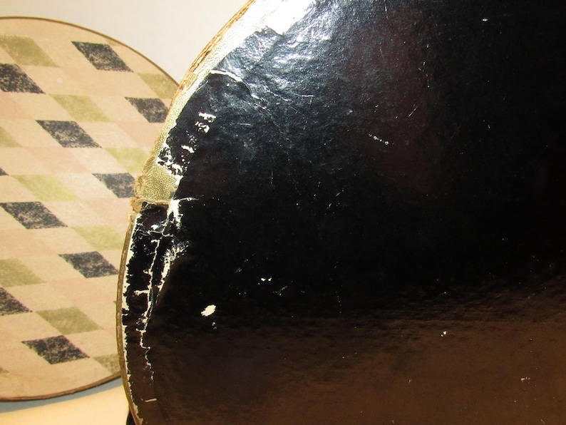 May include: Close-up of two round vintage hat boxes. One box is black with a glossy finish, showing signs of wear and tear. The other box has a cream and tan diamond pattern. The boxes are likely made of cardboard.