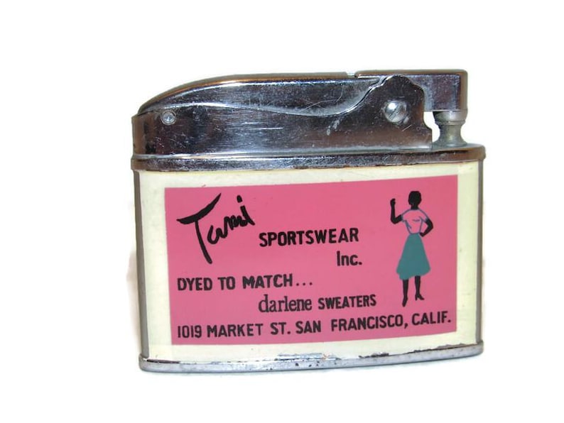 May include: Vintage chrome and cream-colored lighter with a pink label. The label features the text "Tami Sportswear Inc." and a silhouette of a woman. The lighter has a flip-top design.