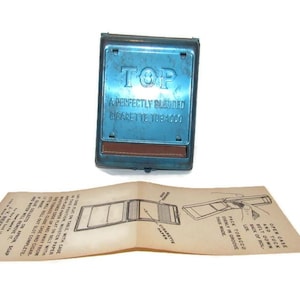 May include: A vintage blue metal cigarette rolling machine with the word "TOP" and the text "A Perfectly Blended Cigarette Tobacco" printed on it. An instruction sheet is below.