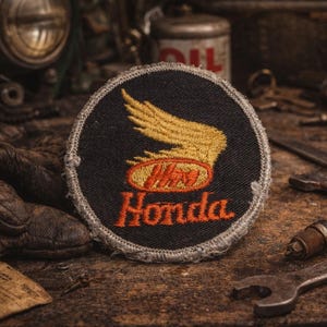 Puede incluir: Parche redondo negro con el logotipo de Honda, con un diseño de ala dorada y texto naranja. El parche está rodeado de herramientas y objetos vintage, lo que sugiere un tema de motocicleta o automoción.