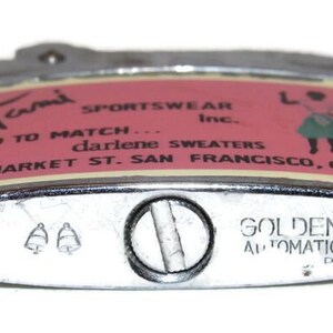 May include: Vintage Golden Bell automatic lighter with a silver finish. The lighter features a pink and cream label with the text "Tami Sportswear Inc." and "Golden Bell Automatic Lighter".