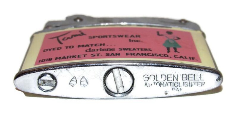 May include: Vintage Golden Bell automatic lighter with a silver finish. The lighter features a pink and cream label with the text "Tami Sportswear Inc." and "Golden Bell Automatic Lighter".