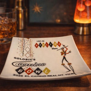 May include: A square, white ceramic plate with a retro design. The plate features the text "GOLOBIC'S Camino BOWL" and "2025 EL CAMINO REAL MT. VIEW". It also has a graphic of a person holding a stick and a series of diamond shapes.