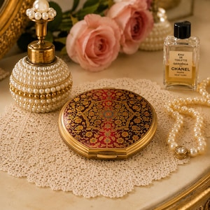 May include: A round, gold-toned compact mirror featuring a detailed, intricate floral pattern in shades of deep red, gold, and black. The compact is positioned on a white lace doily alongside a pearl-adorned perfume bottle and a Chanel perfume bottle.