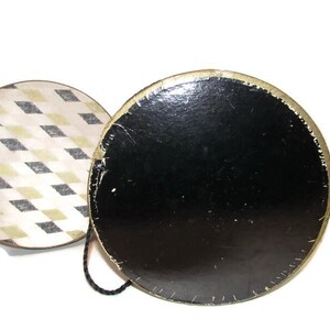May include: A vintage round hat box with a black base and a lid featuring a diamond pattern in cream, black, and gold. The box has a gold trim and a black cord handle.