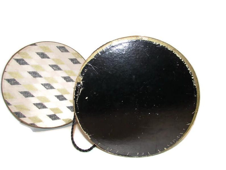 May include: A vintage round hat box with a black base and a lid featuring a diamond pattern in cream, black, and gold. The box has a gold trim and a black cord handle.