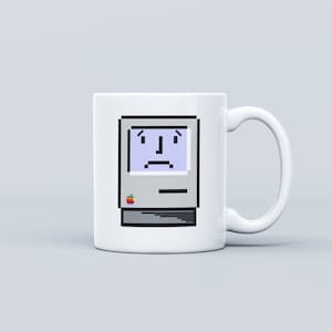 May include: White ceramic mug with a pixelated image of a vintage Macintosh computer with a sad face on the screen. The computer has a rainbow apple logo on the front.
